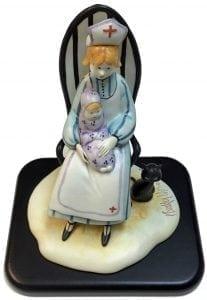 Nurse Figurine titled Loving Care is a P Buckley Moss figurine featuring a nurse holding a baby and giving comfort. Colors of lavender, blue, cream, golden for hair and black base and tiny cat. 
